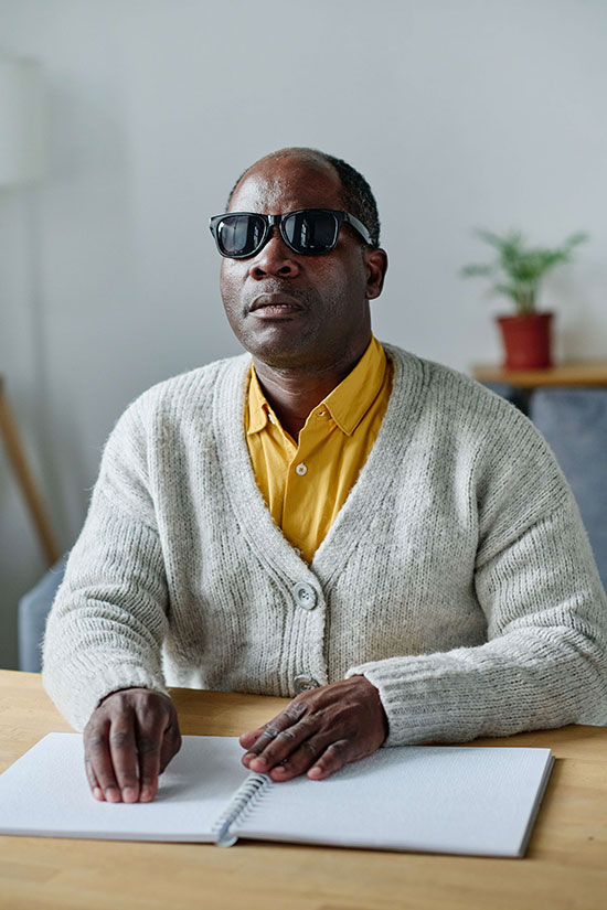 A visually impaired man in sunglasses reads braille at a table.
