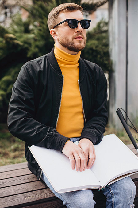 A visually impaired man in sunglasses reads braille book on a bench.