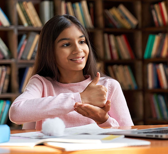 A girl using signs language saying thank you in a library with books and a laptop nearby.
